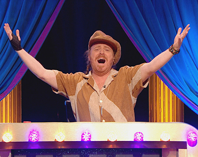 Man singing with arms outstretched on a game show set.