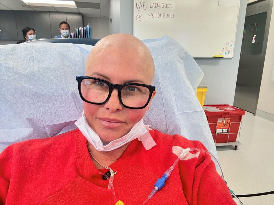 Nicole Eggert, bald and wearing glasses, in a hospital room during cancer treatment.