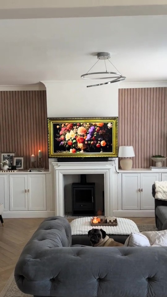 Living room with a fireplace, TV displaying a floral painting, and a pug on a sofa.