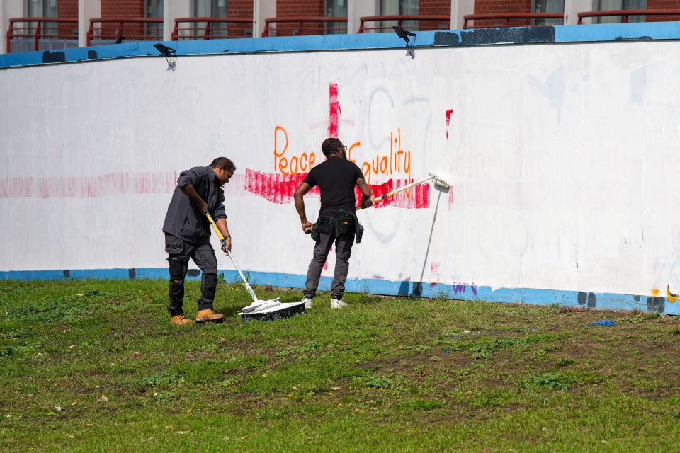 Two maintenance workers painting over graffiti on a wall.