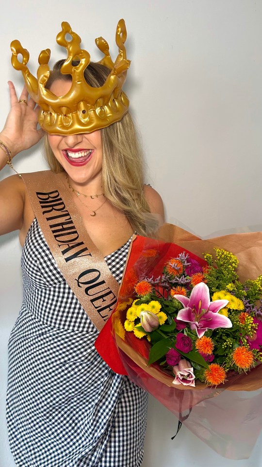 Woman wearing a birthday queen sash and inflatable crown holding a bouquet of flowers.
