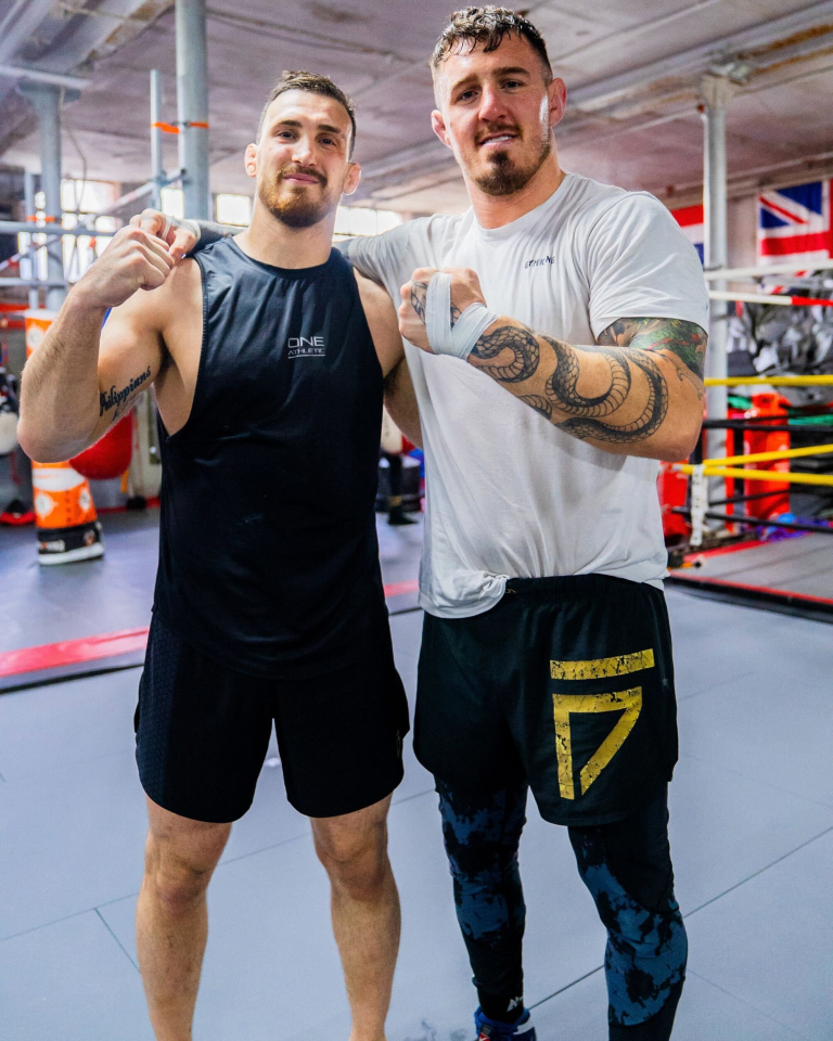 Two men in athletic wear stand together in a boxing gym.