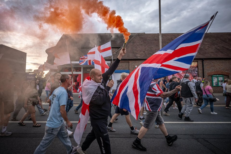 Anti-migrant protesters march through Epping, UK, carrying Union Jack flags and a lit flare.