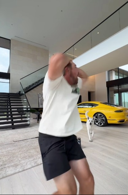 Man jumping in a large house with a yellow car and a white dog.
