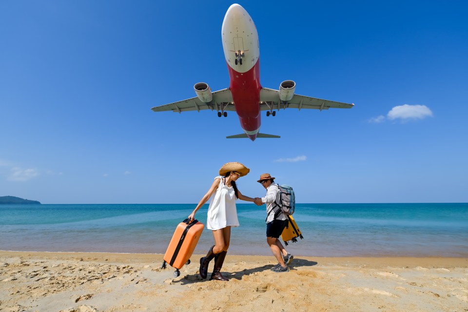 Couple walking on a beach with luggage, airplane landing overhead.