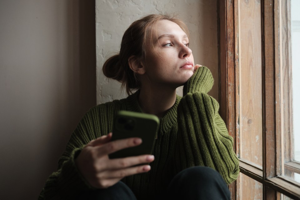 Sad young woman looking out a window while holding her phone.