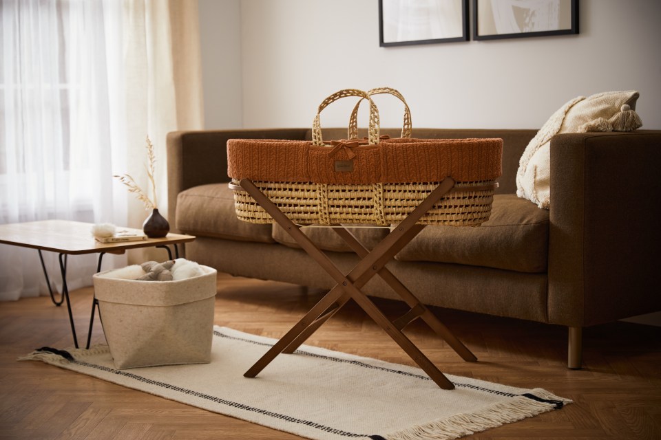 Wicker baby bassinet with terracotta knit liner on wooden stand.