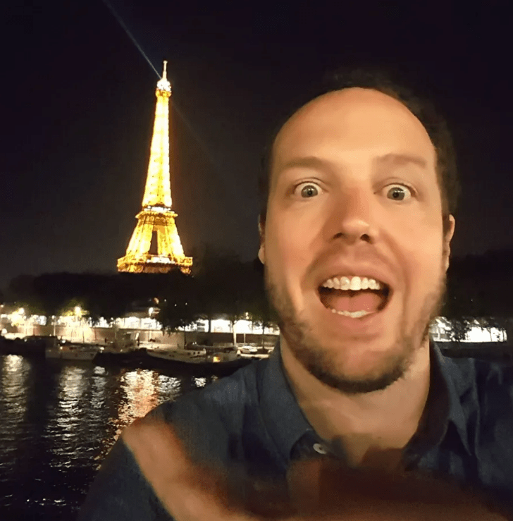 Selfie of a surprised man in front of the Eiffel Tower at night.