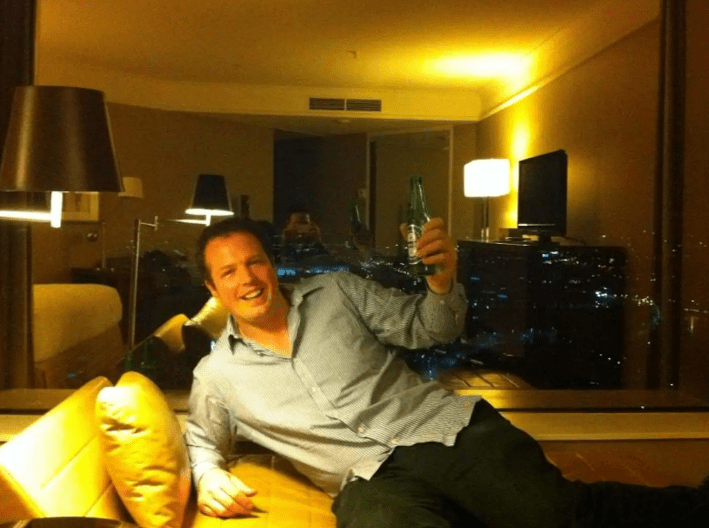 Man relaxing on a couch in a hotel room, raising a bottle in a toast.