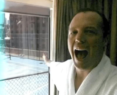 Man in bathrobe excitedly points out a window at a tennis court.