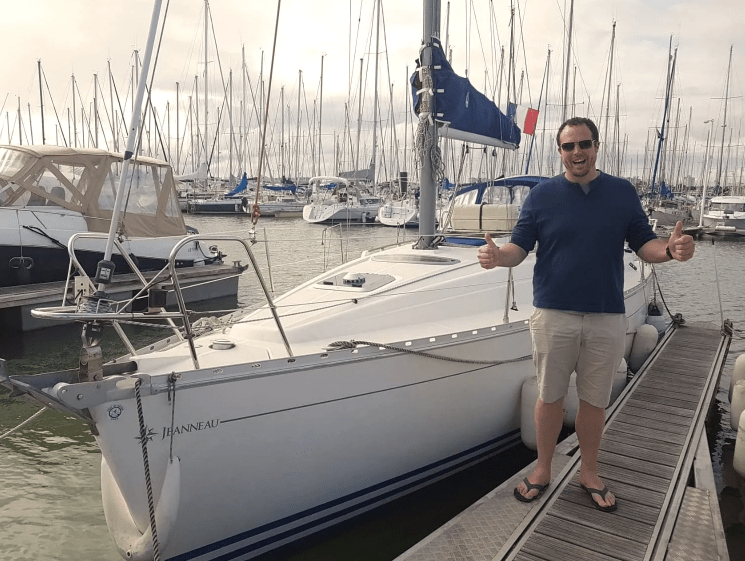 Man giving thumbs up in front of sailboat at marina.