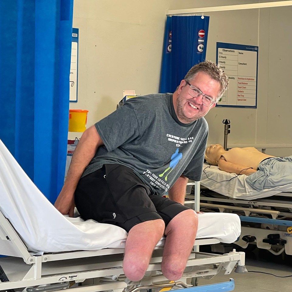 A smiling man with leg amputations sits on a hospital bed.