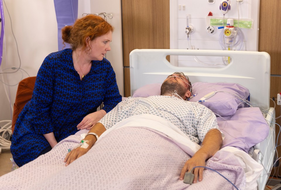 A woman sits beside a man lying in a hospital bed.