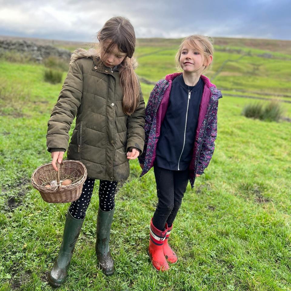 Two girls in rain boots carrying a basket of eggs in a field.