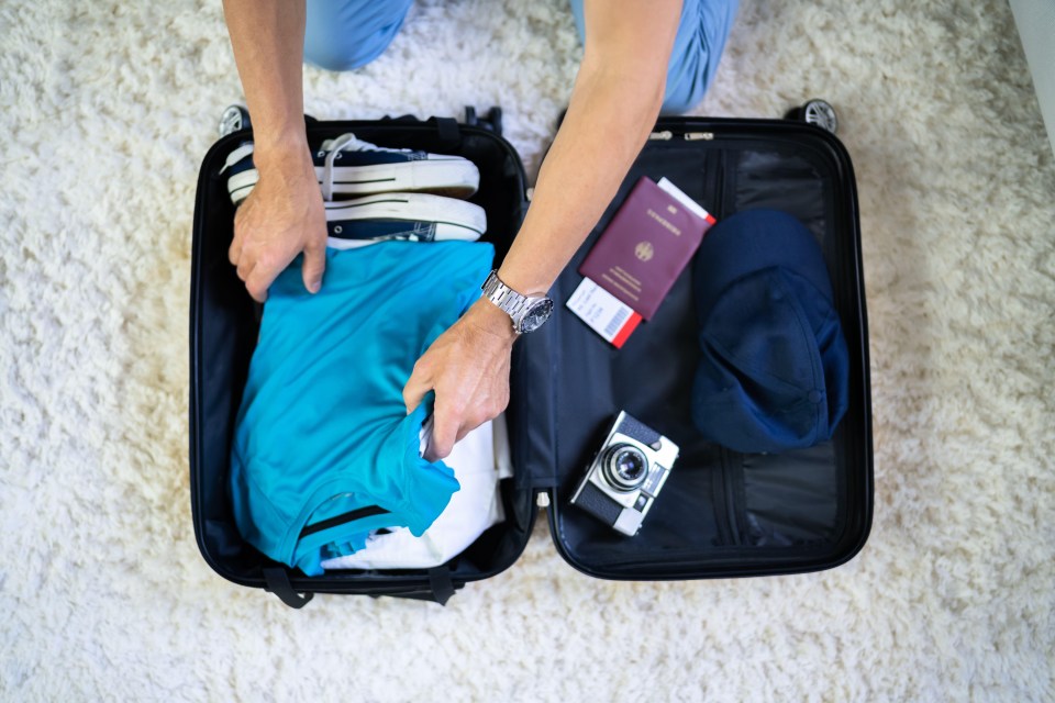 Person packing a suitcase with clothes, shoes, a passport, and a hat.