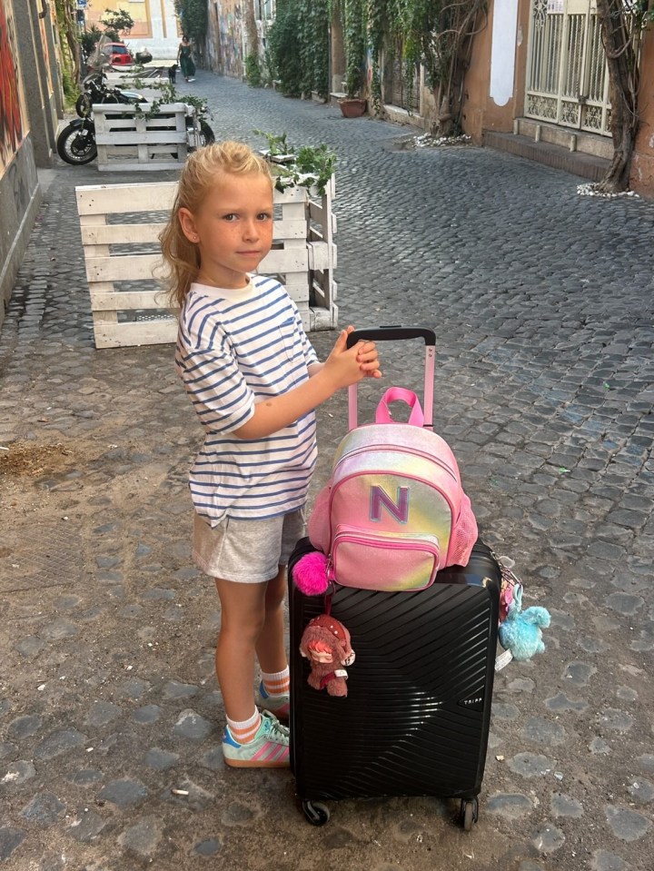 Girl standing with a rolling suitcase and backpack.