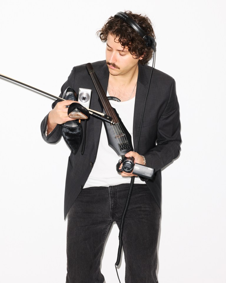 Kozlow, Brooklyn-based artist, holding an electric violin, bow, camera, and shoe.