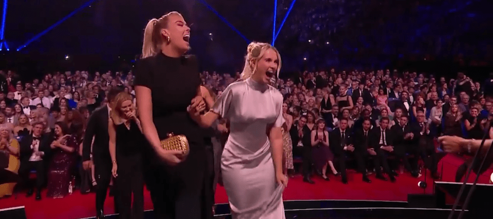 Two women laughing and holding hands while accepting an award in front of a large audience.