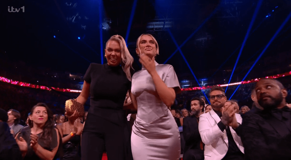 Two women walking and smiling, surrounded by an audience applauding.