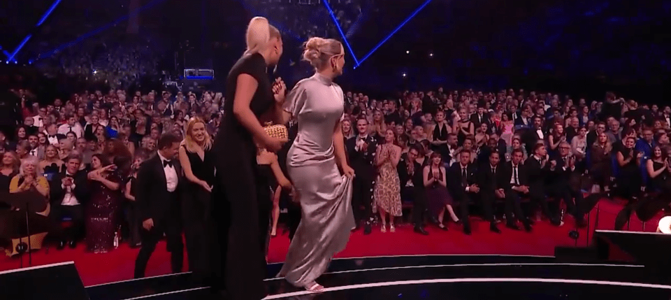 Two women walking across a stage to an applauding audience.