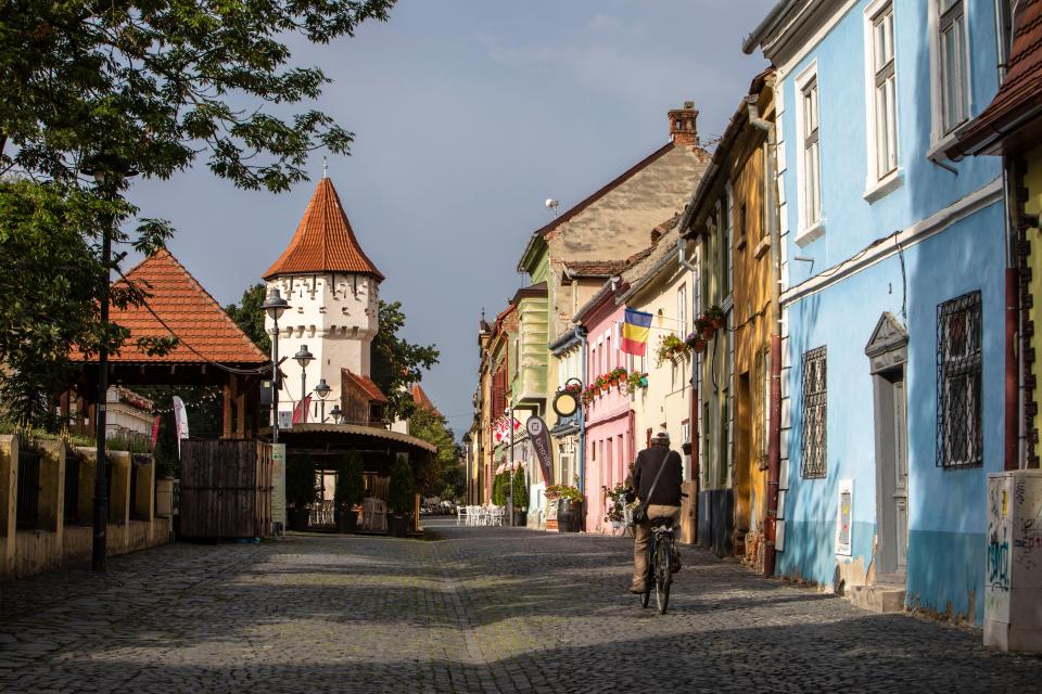 Person biking down a cobblestone street in Sibiu, Romania, past colorful buildings and a medieval tower.