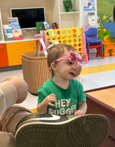 Toddler wearing pink sunglasses sits on the floor playing with a large shoe.