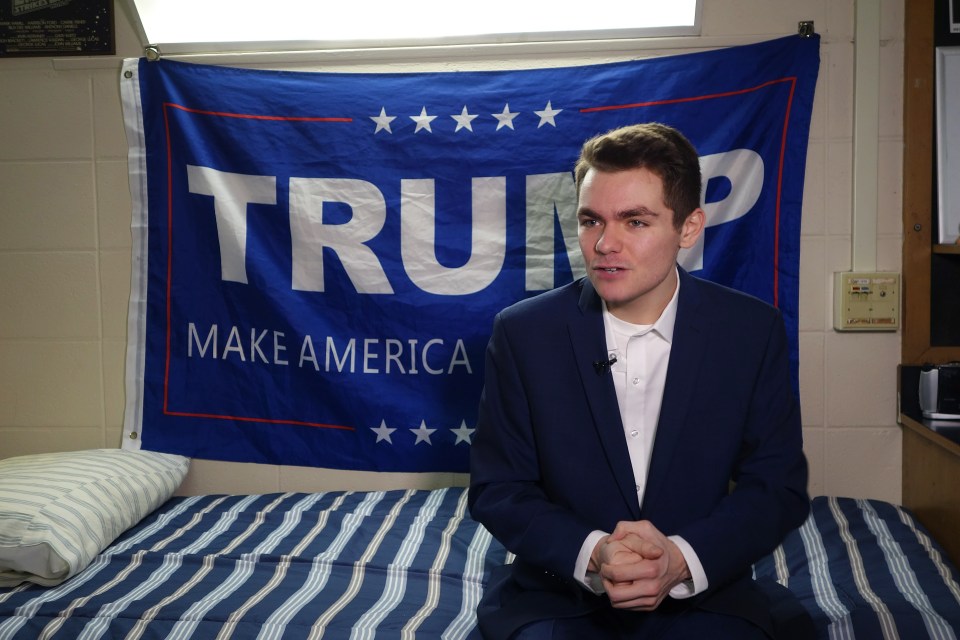 A young man in a suit sits on a bed in a dorm room, a Trump campaign flag behind him.