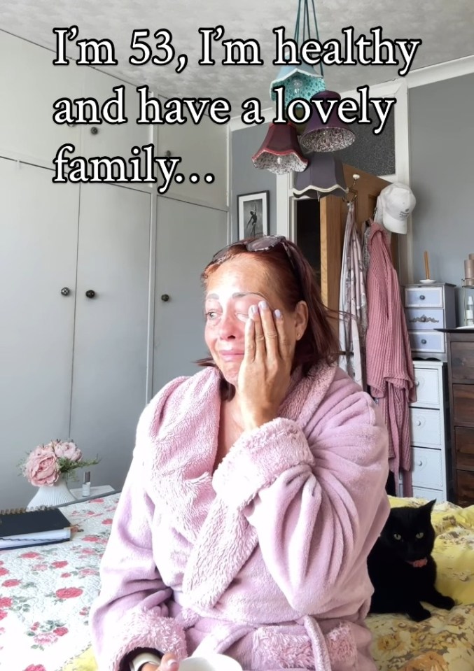 A 53-year-old woman in a pink robe sits on her bed with a black cat; text overlay reads, "I'm 53, I'm healthy and have a lovely family..."
