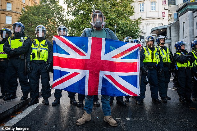 A protester in a gas mask holds a Union Jack flag in front of a row of police officers in combat helmets