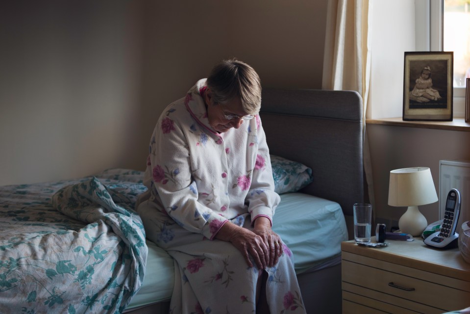 A sad senior woman sits on the side of her bed in her pajamas.