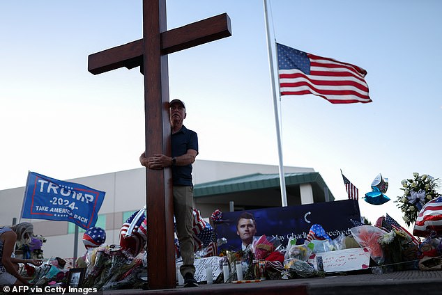 The stars and stripes flag flies at half-mast as a man carries a large cross near a makeshift memorial for Charlie Kirk outside of the Turning Point USA headquarters in Phoenix, Arizona