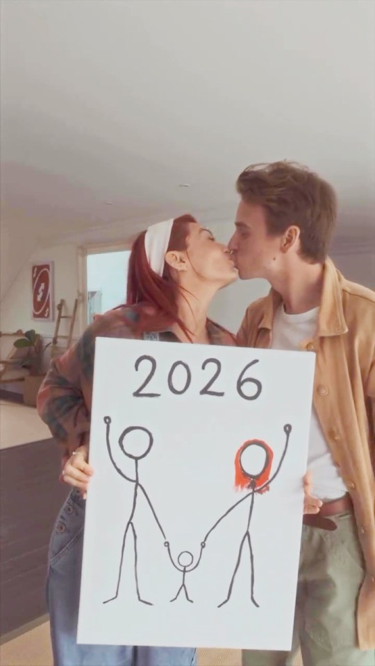 Dianne Buswell and Joe Sugg kissing, holding a sign that says "2026" and depicts a stick figure family.