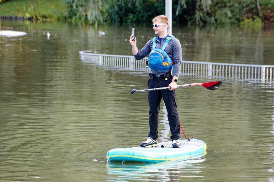 Man paddleboarding in floodwaters, taking a picture with his phone.