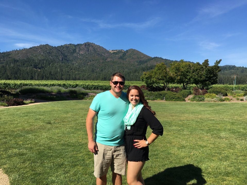 Couple standing on a grassy lawn in front of a vineyard and mountains.