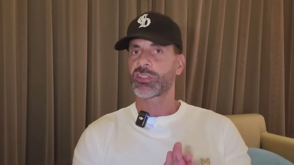 Man wearing a white shirt and black baseball cap with a "SD" logo, speaking with a microphone clipped to his shirt.