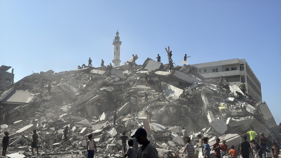 People inspect the destruction of a building, which is now a pile of rubble with a minaret in the background.