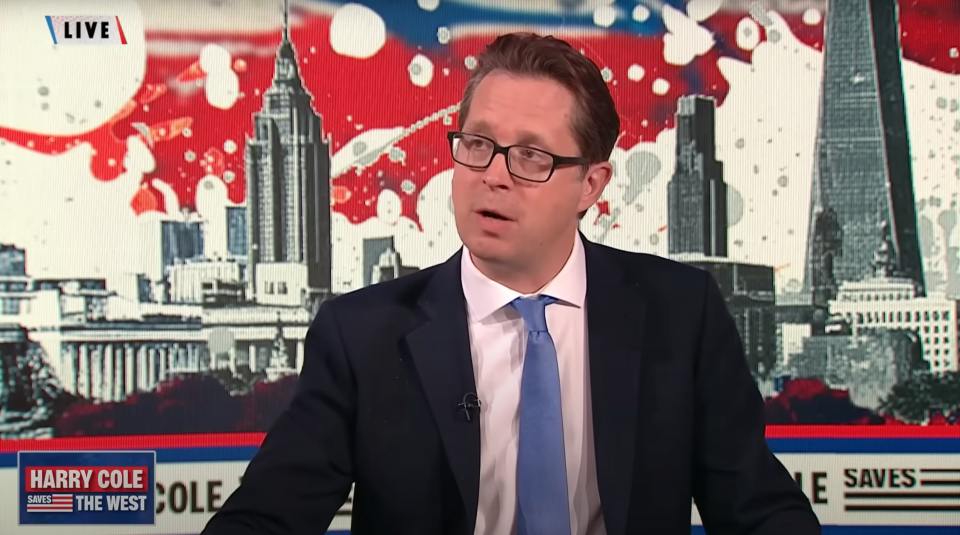 A man in a suit and glasses on a news program with a cityscape background.