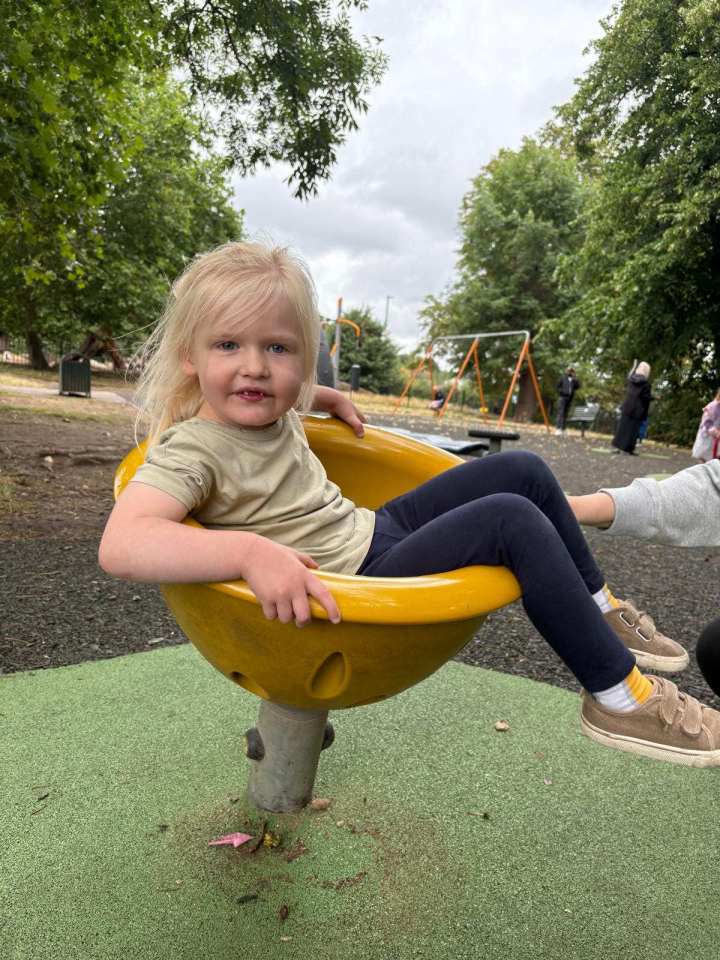 Rosie Bridge, a young girl with blonde hair and blue eyes, sitting in a yellow spinning bowl at a playground.