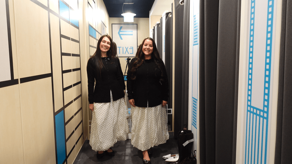 Two women stand in a hallway with changing rooms to the right and an exit sign behind them.