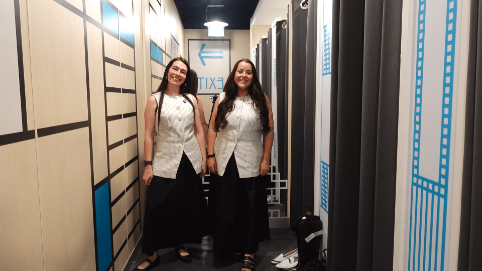 Two women in a narrow room with Mondrian-style walls on one side and black curtains on the other, an exit sign at the end of the room.
