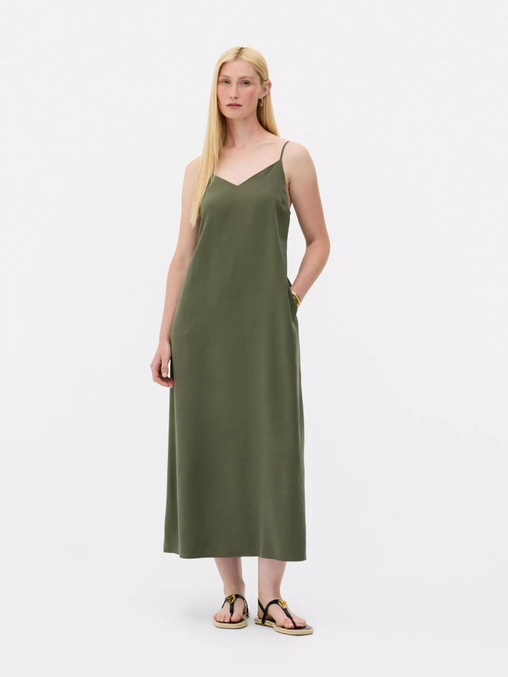 Woman with long blonde hair wearing a green midi slip dress and black flat sandals.