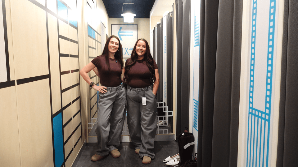Two women wearing brown shirts and gray pants in a hallway.