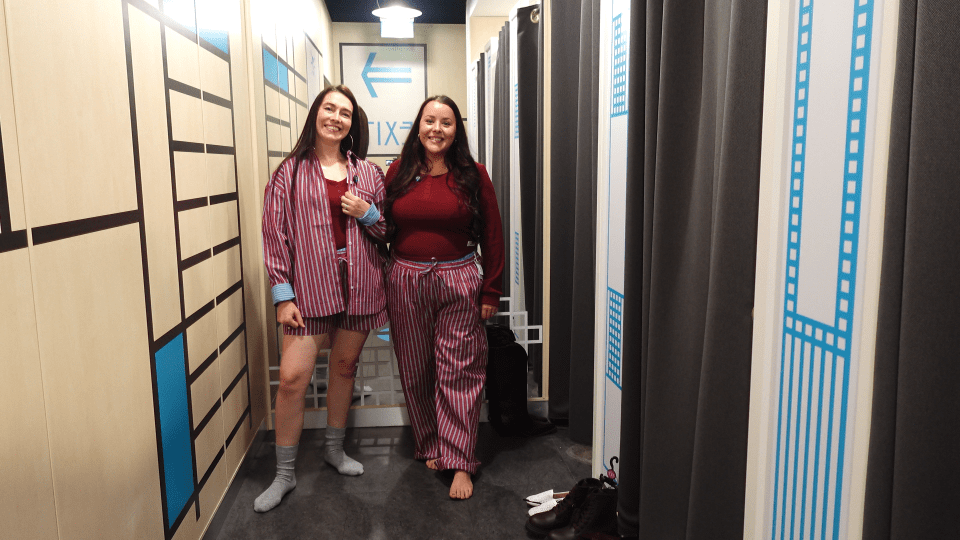 Two women dressed in striped pajamas with one wearing shorts and the other wearing pants, standing in a hallway.