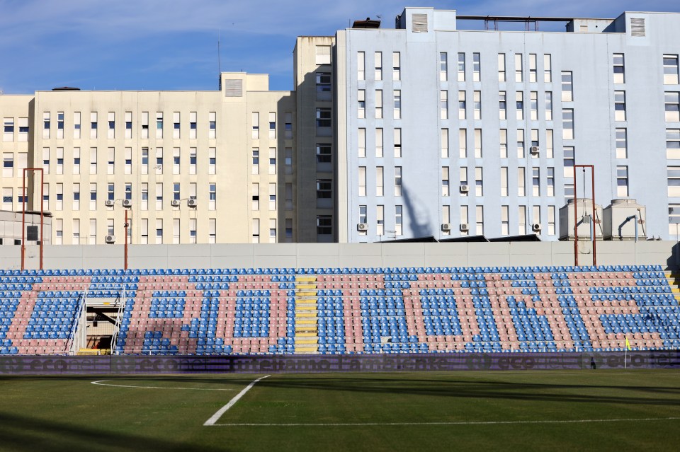 General view of the Stadio Ezio Scida in Crotone, Italy, showing blue and pink stadium seating with buildings in the background.