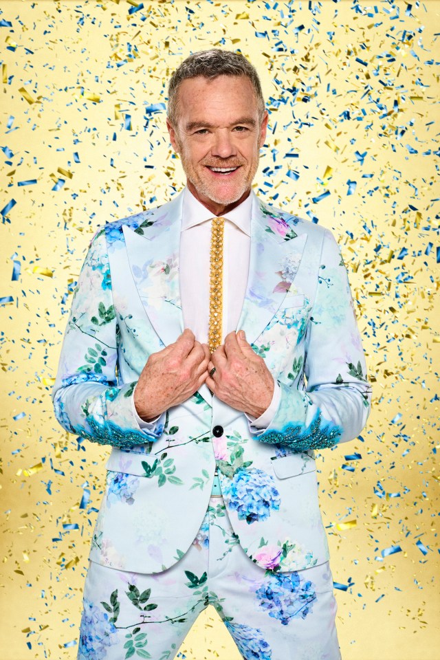 Stefan Dennis in a pale blue floral suit with a decorative gold tie, smiling against a confetti-filled yellow background.