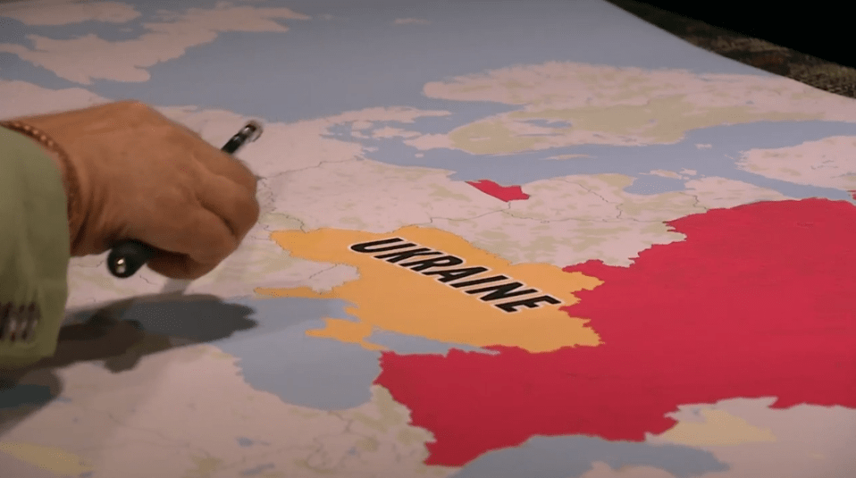 A hand with a pen pointing to a map of Ukraine colored yellow, with surrounding countries in red.