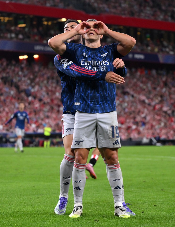 Leandro Trossard celebrates scoring by holding his hands to his eyes to form the shape of a heart, with Gabriel Martinelli hugging him from behind.