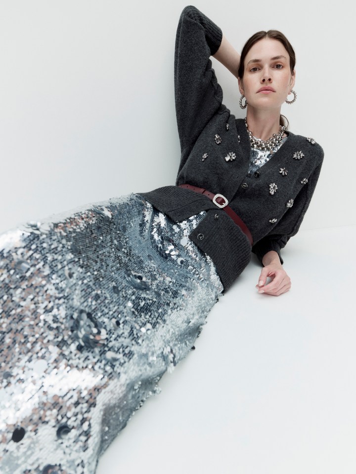 A model wears a gray cardigan with jewel accents, a silver sequin top, a silver sequin skirt, and a thin brown belt.