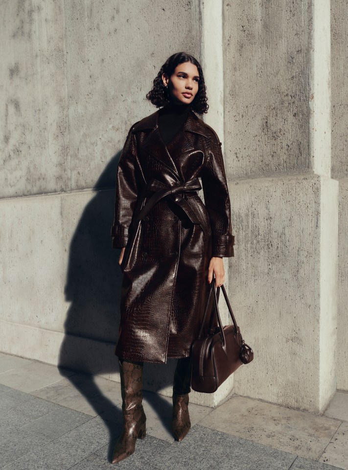 A model in the M&S Autumn Winter 2025 collection wearing a brown crocodile-patterned trench coat, dark turtleneck, snakeskin boots, and holding a brown leather bag.