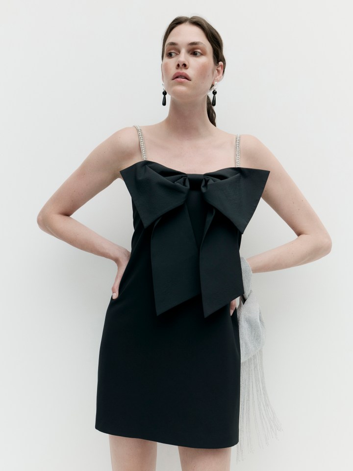 Woman models a black mini-dress with a large bow detail and crystal-embellished straps for the M&S Autumn Winter 2025 collection.